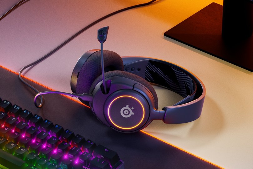 An image of Arctis Nova headset laying on the desk, accompanied with glowing keyboard and mouse.